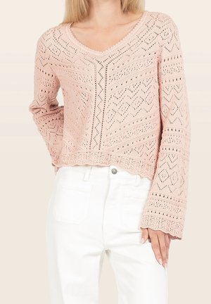 Mujer con un suéter de punto de ganchillo de color rosa claro con mangas largas y pantalones blancos de cintura alta, con la mano apoyada en el muslo.