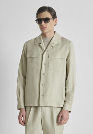 Homme portant une chemise beige boutonnée et un pantalon assorti avec des lunettes de soleil foncées sur un fond clair uni.