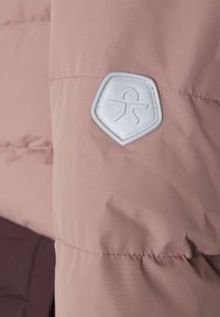 Tessuto della giacca imbottita rosa chiaro con una texture liscia. Presenta una toppa grigia a forma di diamante sulla manica sinistra e dettagli cuciti.