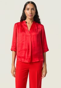 Blusa de satén roja con escote en V, mangas tres cuartos y corte holgado. Textura suave, detalles mínimos, combinada con pantalones rojos.