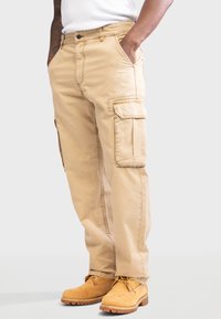 Tan cargobroek met zijzakken, losse pasvorm, duurzaam materiaal. G gedragen met gele werkschoenen en een wit shirt.