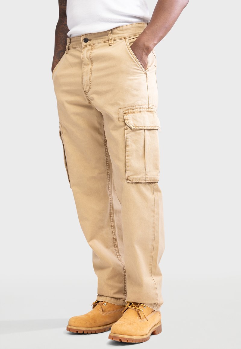 Tan cargobroek met zijzakken, losse pasvorm, duurzaam materiaal. G gedragen met gele werkschoenen en een wit shirt.