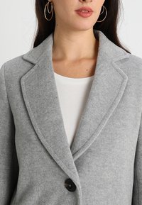 Grijze wollen blazer met een nietched kraag, sluiting met twee knopen en een zachte textuur. Draagt over een wit shirt en gecombineerd met grote oorringen.