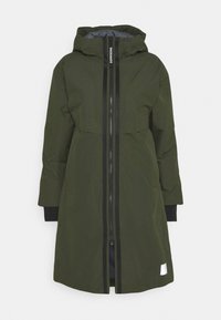 Vihreä takki, joka on valmistettu sileästä, vettä hylkivästä materiaalista ja jossa on vetoketjusuljenta sekä huppu. Varustettu kontrastivärisillä mustilla vetoketjuilla ja hihansuilla.