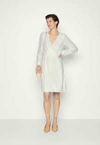 Vestido branco de manga longa, tecido texturizado e cintura ajustada; acompanhado de sandálias beige de tiras e uma pequena bolsa beige.