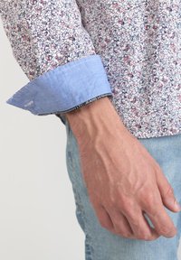 Camicia con motivo floreale bianco e pastello, caratterizzata da un polsino azzurro chiaro a contrasto e dettagli di cucitura visibili. Mano in denim.