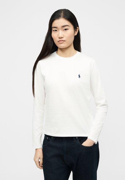 Νεαρή γυναίκα με μακριά σκουρόχρωμα μαλλιά, φορώντας λευκό μακρυμάνικο πουκάμισο με μικρό ναυτικό λογότυπο στο στήθος και σκούρο τζιν, στέκεται όρθια.