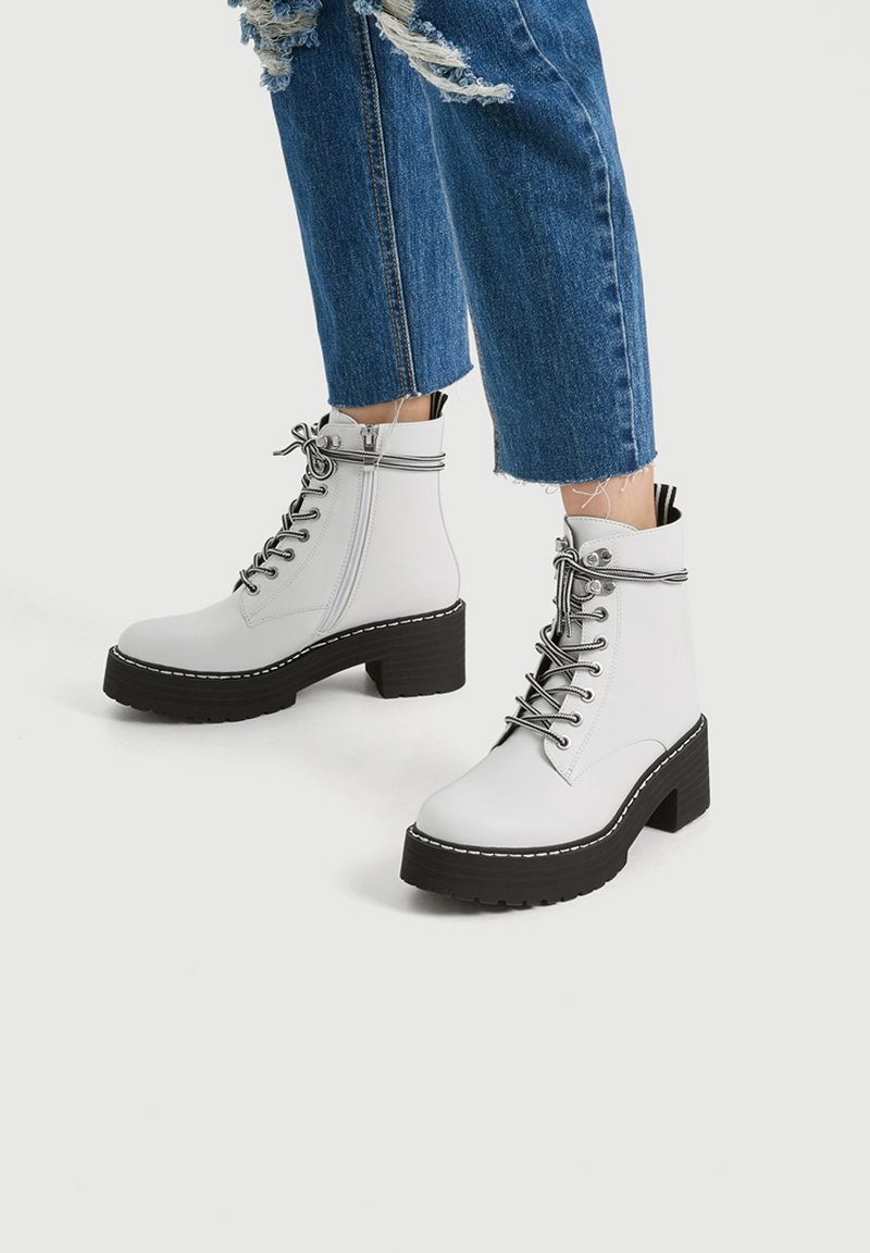 Bottes blanches à lacets avec semelles épaisses noires portées avec un jean bleu déchiré et court sur fond blanc.