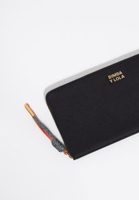 Cartera negra con el logo dorado "Bimba y Lola". Presenta un material texturizado y un cierre con un pompón multicolor hecho de gris, rojo y dorado.