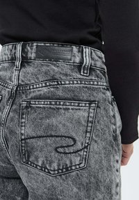 Personne portant un haut noir et un jean en denim gris décoloré avec des coutures sur les poches arrières et une étiquette de marque en cuir noir sur la taille.