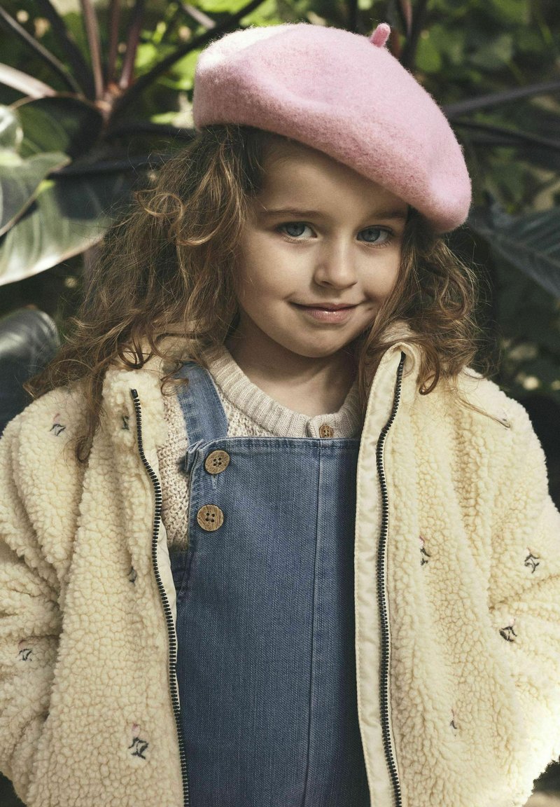 Kind met een roze vilten baret, een zachte crèmekleurige sherpa-jas met bloemenborduursel, en blauwe denim tuinbroek met houten knopen.