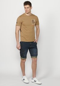T-shirt en coton marron avec un imprimé graphique sur la poitrine, associé à un short en denim cargo. Le short a des ourlets retroussés et des poches. Baskets blanches.