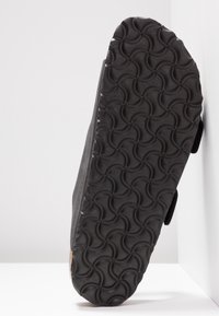 Chaussure noire vue de dessous, montrant une semelle sombre avec un motif géométrique circulaire répétitif sur fond blanc.