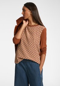 Maglia marrone a maniche lunghe con parte anteriore crema a fantasia e maniche marroni uni. Abbinata a pantaloni in denim a gamba larga di colore blu scuro.