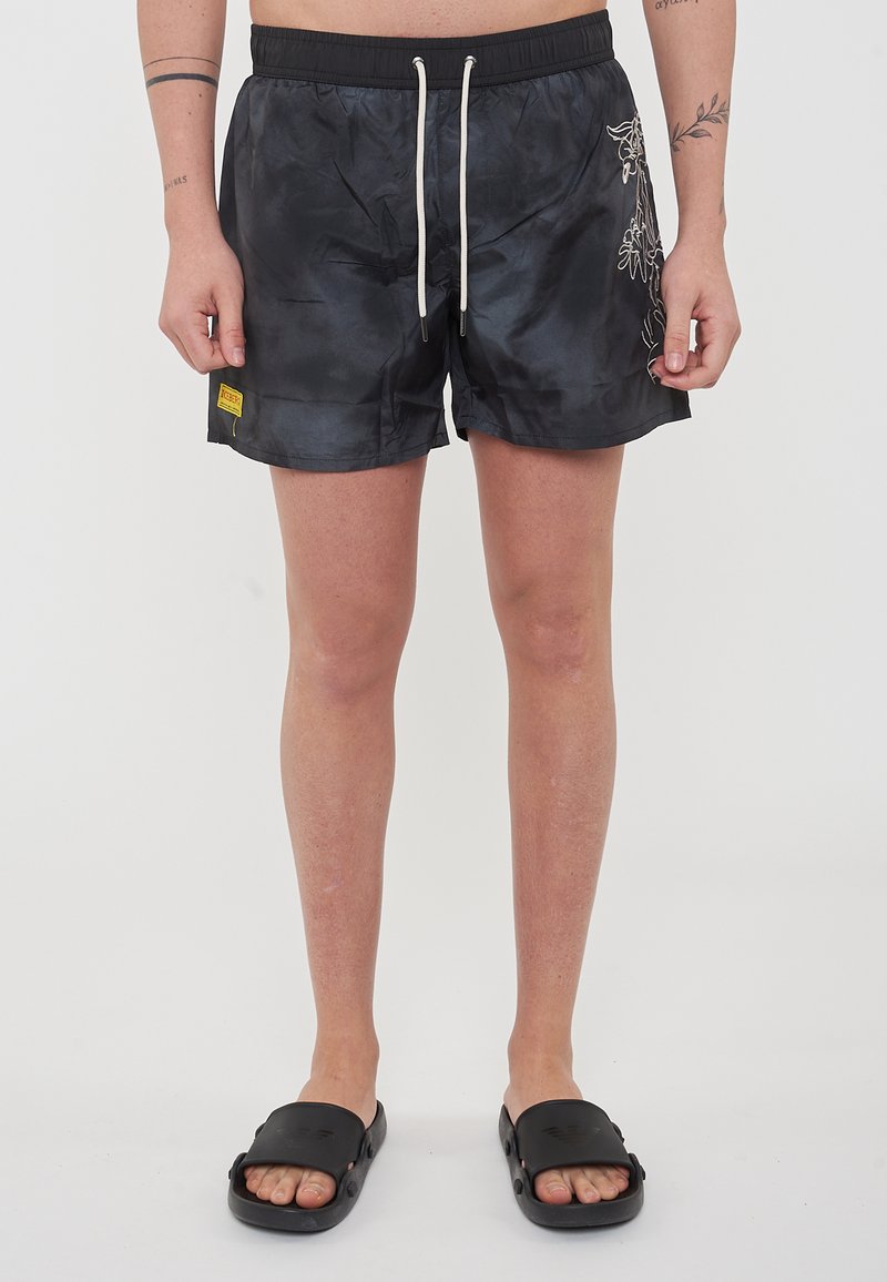 Pantaloni da bagno neri con cintura elastica, cordino bianco, tasche laterali e design floreale. Indossati con sandali neri a scivolo.