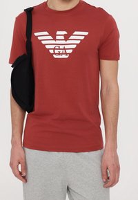 Czerwona koszulka z krótkim rękawem z białym logiem orła i paskami. Zestawiona z szarymi spodniami dresowymi i czarną torbą. Materiał bawełniany.