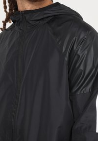 Czarna, lekka kurtka o gładkiej, błyszczącej fakturze, z kapturem i zapięciem na zamek. Posiada rękawy raglanowe dla swobody ruchów.