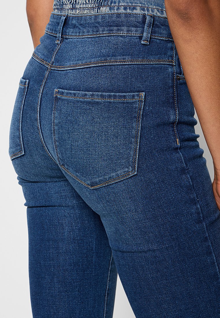 Gros plan sur une personne portant un jean en denim bleu foncé taille haute avec des poches arrière et des coutures visibles.