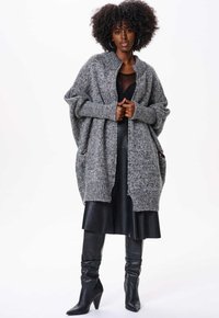Gilet en tricot gris avec une texture épaisse, à l'avant ouvert et avec des poches latérales, porté sur une robe noire et des bottes noires à talons hauts.