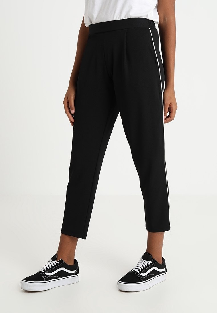Pantalons noirs coupés avec des rayures blanches sur les côtés, présentant une texture lisse et une taille élastique. Assortis avec des baskets noires et blanches.