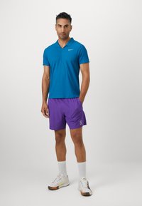 Férfi kék pólóing gallérral, lila sportnadrággal párosítva. Fehér sportcipők színes akcentusokkal és fehér bokazokni.