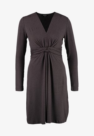 Femme portant une robe gris foncé à manches longues et longueur genoux avec un nœud sur le devant, des collants noirs, des talons noirs, et tenant un sac à main noir avec une bandoulière en chaîne.