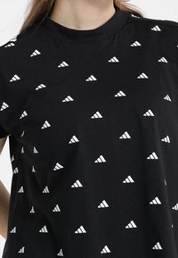 Černé tričko s krátkými rukávy a opakujícím se bílým trojúhelníkovým vzorem. Látka působí měkce, s kulatým výstřihem a volným střihem.