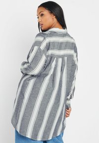 Camisa de manga larga con rayas verticales en gris y blanco, dobladillo redondeado y detalles de botones en las mangas. Tela texturada con un corte relajado.