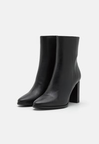 Botas de tobillo de cuero negro con un diseño de punta afilada, que cuentan con un tacón grueso y una textura suave. Estilo y silueta minimalistas.