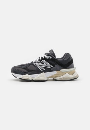 New Balance-sneaker i sort og gråt netmateriale med ruskindsdetaljer, hvide snørebånd og en beige gummisål. Har en fremtrædende logo på siden.