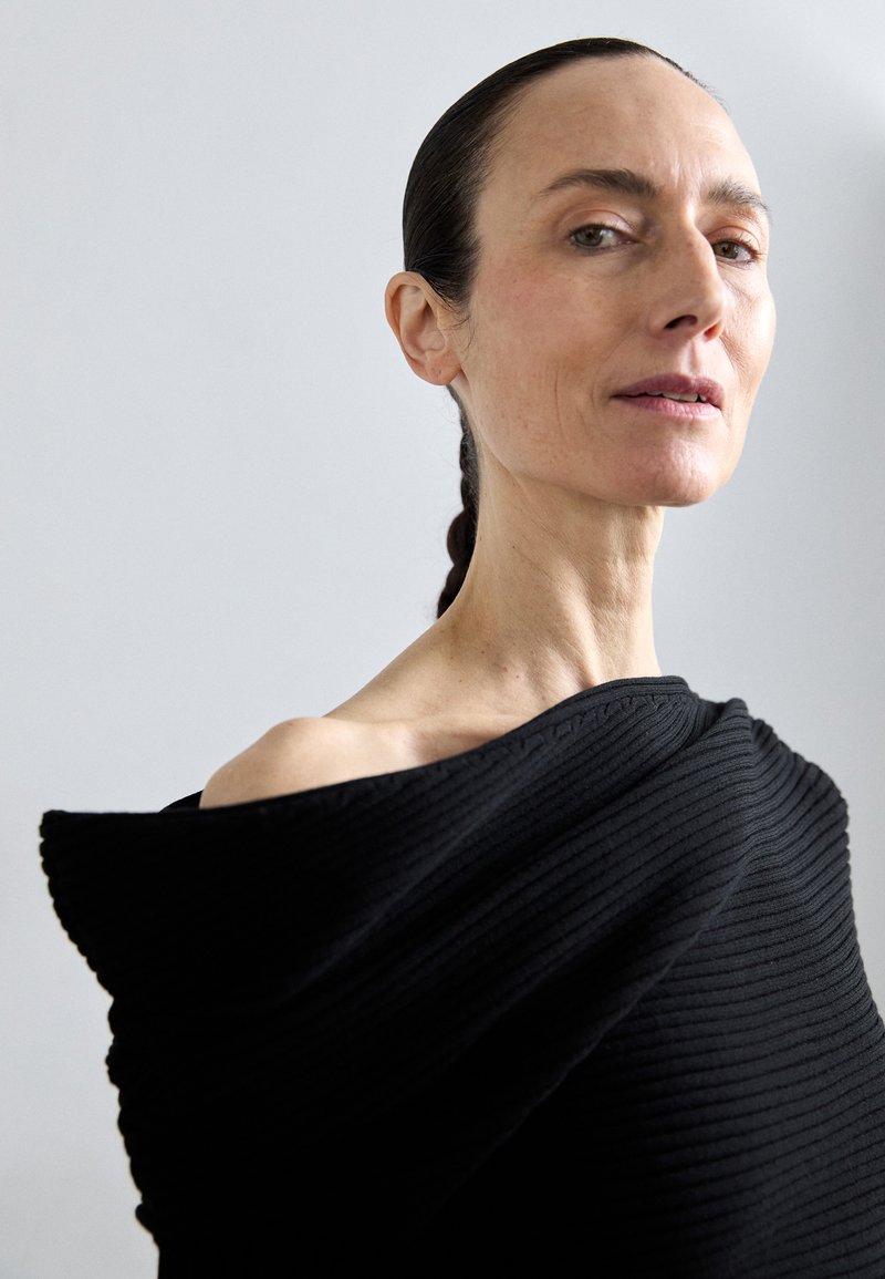  Haut noir côtelé à épaules dénudées avec une finition texturée. Le tissu drape élégamment, mettant en valeur le décolleté et les clavicules.