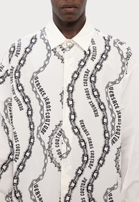 Camisa blanca con un patrón de cadenas y texto en negro. Diseño de botones con cuello y tejido texturizado, que muestra elementos gráficos audaces.