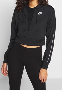 Kort zwart uitgevoerde jack met rits, gemaakt van gladde stof. Het heeft een capuchon, witte strepen aan de zijkanten en een klein wit logo op de borst.