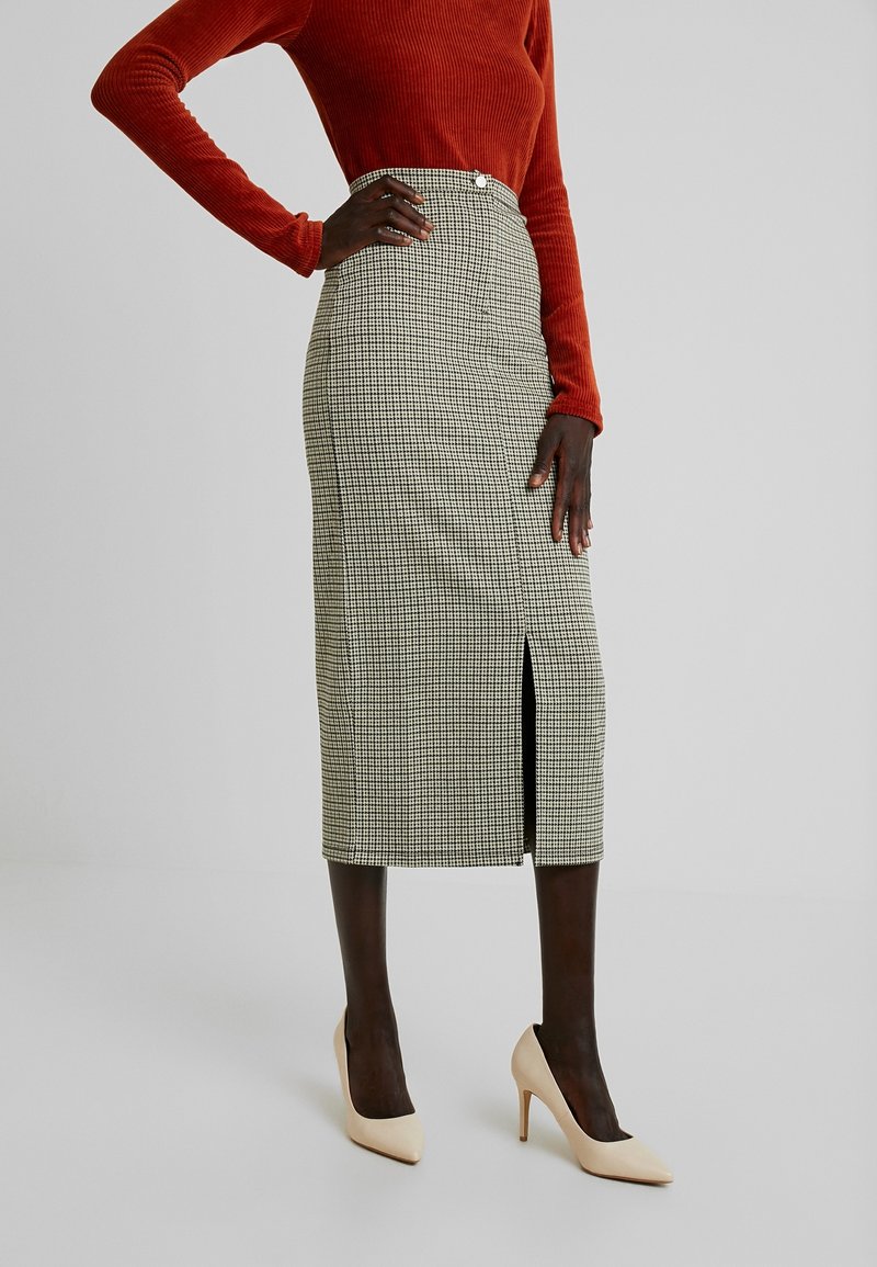 Mannequin portant une jupe midi taille haute à motif pied-de-poule avec une fente devant, un top à manches longues couleur rouille, et des chaussures à talons beiges sur fond gris.