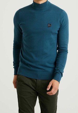 Mężczyzna w turkusowym swetrze z golfem z małym czarnym logo na piersi oraz ciemnooliwkowych spodniach, stojący na jednolitym tle.