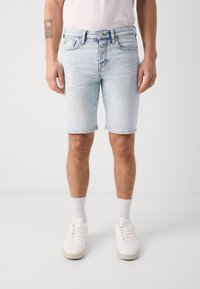 Shorts en denim bleu clair avec une coupe droite, ourlets retroussés, cinq poches et une fermeture à bouton. Associés à des baskets blanches et des chaussettes.