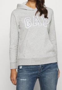 Sudadera de color gris claro hecha de tejido suave, con un bolsillo canguro frontal y el logotipo "GAP" en letras blancas contorneadas. Puños y dobladillo de canalé.
