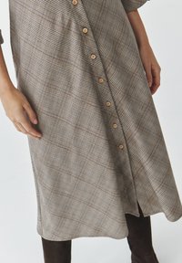 Abito camicia a quadri marrone realizzato in tessuto testurizzato, caratterizzato da bottoni in legno, un orlo curvo e diverse fantasie a quadri.