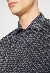 Černá košilové s krátkým rukávem s bílým geometrickým vzorem. Obsahuje klasický límec a dva šedé knoflíky na předním zapínání.