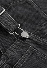 Primo piano di una bretella di salopette in denim scuro attaccata a una fibbia metallica con dettagli di cucitura visibili sul tessuto e sul bordo della tasca.