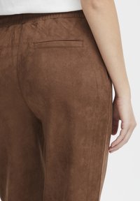 Brown suede pants with a smooth texture, featuring back pockets and an elastic waistband. The image shows the back view and portion of a hand.