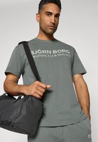 Pelēks kokvilnas T-krekls ar uzrakstu "BJÖRN BORG TRAINING CLUB MONACO", kas izdrukāts baltā krāsā. Tur rokās melnu sporta somu ar redzamu rāvējslēdzēju un rokturi.