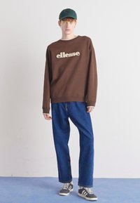 Sudadera marrón con el logo de "ellesse", combinada con jeans rectos azules y zapatillas grises. Modelo usando una gorra verde. Fondo simple.