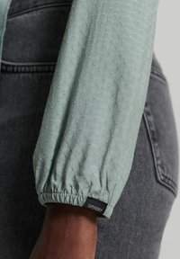 Jasnozielona, pikowana rękawica z marszczonym mankietem, z delikatną teksturą i czarną etykietą "SUPERDRY". Tło stanowi szary dżins.