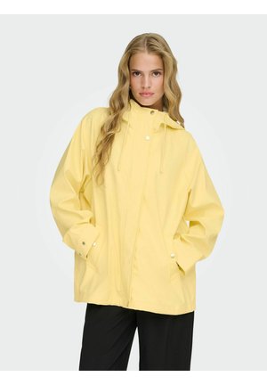 Frau mit langen welligen blonden Haaren, die eine lockere gelbe Regenjacke und schwarze Hosen trägt und mit den Händen in den Taschen steht.