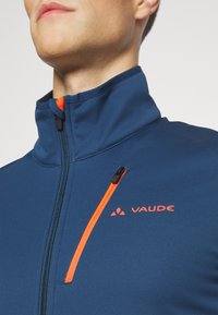 Tamsiai mėlyna užtraukiama striukė su aukštu apykakle, puikuojanti oranžiniu užtrauktuku ir logotipu. Lygus paviršius su prigludusia forma.