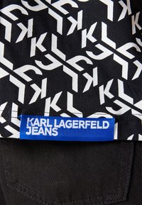 Svart denim med ett vitt geometriskt mönster som har "KL"-logotyper. En blå etikett med texten "KARL LAGERFELD JEANS" finns längs fållen.