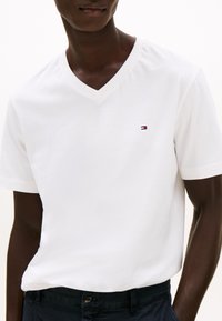 Mann iført hvit V-hals T-skjorte med liten rød, hvit og blå logo på venstre bryst, kombinert med mørke bukser, mot en ensfarget bakgrunn.