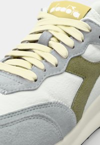 Grey and white sneaker with yellow laces, suede accents, and a green logo. Features stitched detailing and a cushioned sole.