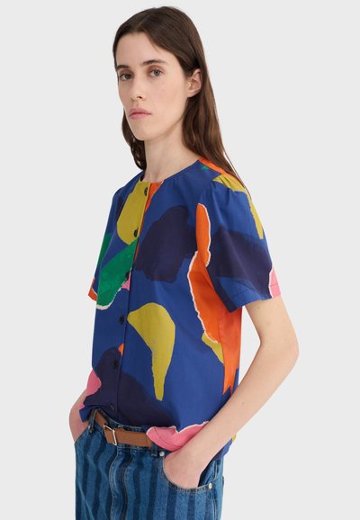 Femme aux longs cheveux bruns portant un chemisier à manches courtes à imprimé abstrait coloré et un jean rayé bleu avec une ceinture marron, les mains dans les poches.